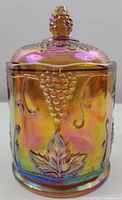 Front view of vintage Indiana Glass Harvest Grape jar showing grape cluster and leaf embossed design on iridescent marigold glass surface, lid and grape cluster handle visible on top.