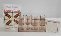 Clear acrylic canapé set with six distinct compartments in different shapes and sizes including cylindrical and rectangular shapes, original colorful printed instruction booklet titled Phyllis Gordon's Short Cuts Canapés the Easy Way, and original packaging box.