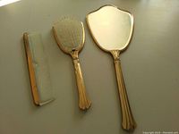 Full view of gold plated hand mirror, hairbrush with white bristles and comb showing slight wear and rust on the mirror surface.