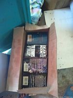 Photo showing box containing several paperback novels with visible titles by Dean Koontz and Stephen King, stacked lying flat.