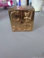 Front side of brass baby block coin bank showing embossed letters C and D with images of a cat and a horse, and a lock mechanism.