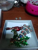 Five brooches including a red rose, flower cluster with rhinestones, and holly leaves with berries.