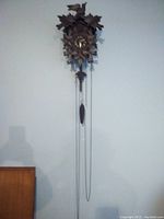 Full view of the Black Forest chalet-style cuckoo clock with carved leaves, bird figure on top, and long pendulum and weights.