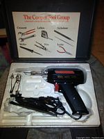 Open black plastic carrying case revealing black pistol-grip soldering iron, power cord, two metal tool attachments, and a printed guide listing tool brand names on case interior lid.
