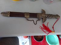 Closed view of the decorative dagger with sheath, showing detailed bronze-colored fittings and attached red tassel.