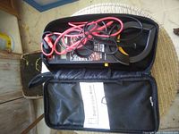 Digital clamp meter placed inside a black carrying case with red and black test probes coiled on top and the user manual in a mesh pocket.