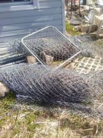 Two rolled metal chain link fences and a detached gate section lying on the ground outdoors against the side of a building.