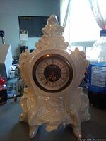 Front and side view of white/cream iridescent mantel shelf clock with ornate design and Roman numeral clock face.