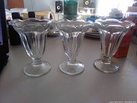 Photo showing three clear glass dessert parfait cups with fluted rounded bowls, scalloped wide rims and pedestal bases placed on a table surface.