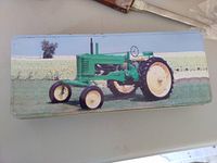 Photo of the rectangular green collectible tin box featuring a green John Deere tractor printed on its lid, placed on a light surface.