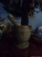 Mortar and pestle set shown on a red surface with various decorative items around it. The mortar is solid marble with natural veining, the pestle rests inside it, matching in material and design.