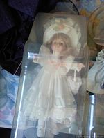 Porcelain doll in plastic case wearing intricate white lace dress and matching bonnet with peach ribbon accents, posed with arms slightly raised.