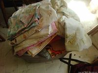 Tote filled with assorted linens in pastel colors and floral prints, placed on tile floor near furniture.