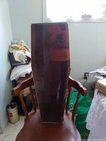 Wooden doll coffin placed on a wooden chair with armrests, showing the coffin's full tapered shape and dark reddish-brown wood finish.