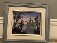 Framed oil painting showing a silhouette of trees by a lake at dusk with a bird in the sky, gray painted wood frame with white inner border.