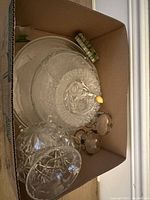 Various glass trays, bowls, and a trinket box with lid along with two small etched glass bowls and a fork with yellow handle in a cardboard box.