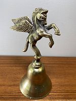 Front view of brass Pegasus bell standing on a wooden surface, showing detailed Pegasus with wings on top of the bell.