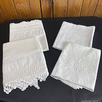 Four white cotton pillowcases arranged in two pairs showing crochet edges on one pair and floral embroidery on another.