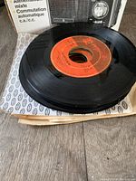 Photo showing stack of vintage 45 RPM vinyl records with orange label on top, packed in box with patterned paper lining