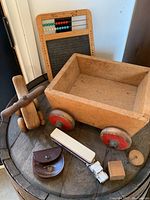 Photo showing vintage wooden wagon, chalkboard with abacus, white stapler, brown pouch, miniature truck, and wooden piece toys on a round table.