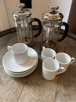 Top view of two glass French presses with chrome tops and black handles, four white espresso cups, and four white saucers arranged on wooden round table.