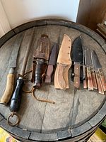 Full view of the collection shown on a wooden barrel surface, includes hunting knives, kitchen knives, and a sharpening tool.