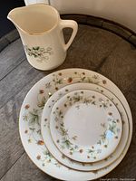 Set of three plates and a ceramic pitcher with floral design and gold rim placed on wooden surface.