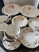 Photo showing a collection of assorted vintage ceramic dishes with various flower patterns, including bowls, cups, and plates with scalloped edges and colored floral prints.