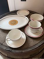 Photo showing oval plate with gold medallion, two cups with red and gold trim, bowls and plates with simple trims arranged on a barrel