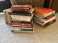 Two stacks of various vintage cookbooks on carpet floor with a wooden barrel in the background, showing a range of titles and conditions.