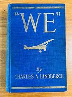 Front cover of the blue hardcover book titled 'WE' with silver embossed details including an airplane illustration and author's name Charles A. Lindbergh.