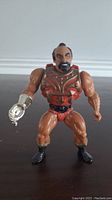 Front view of vintage 1980's He-Man Masters of the Universe Jitsu action figure showing tan muscular body, red and gold armor, silver robotic claw right hand, and facial details.