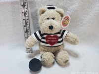 Front view of the Starbucks Bearista teddy bear standing next to a ruler for size reference. The bear is beige, fuzzy, and dressed in a black and white striped shirt with a red heart that reads 'UNCHAIN MY HEART'. The bear wears a small hat.