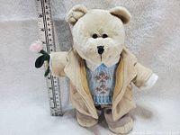 Front view of the teddy bear with measuring ruler showing approximately 9 inches height. Bear wears tan jacket, blue knitted vest, and holds pink flower.