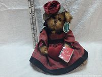 Front view of Bearington teddy bear sitting, dressed in a burgundy and black dress with matching hat, holding a small book, with ruler for size reference.