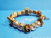 Gold tone stretch bracelet with rhinestone, rose, and bow details on blue background, angled view.