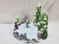 Porcelain figurine hand painted with winter skating pond scene including two skaters, stone wall and snow-covered trees.