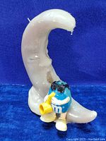 Frontal view of the M&M's blue character sitting on a crescent moon candle while playing a yellow saxophone.