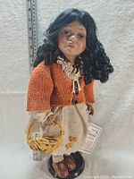 Full doll view showing entire outfit, including orange cardigan, beige skirt with floral detail, brown shoes, and basket accessory. Ruler in background for size reference.
