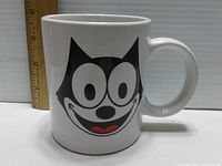 Front view of the white ceramic coffee mug with Felix the Cat face, alongside a ruler for size reference.