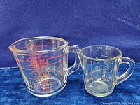 Photo showing the lot of 2 glass measuring cups side by side on a blue fabric background. One cup has red Glassbake markings, the other smaller cup is unmarked with white markings.