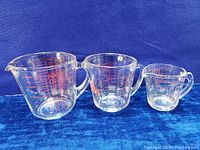 Set of three clear glass measuring cups with red markings and handles, arranged side by side showing size differences and Pyrex logos.