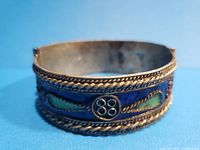 Front view of vintage Moroccan hinged enamel bracelet showing blue, green, and yellow enamel decoration bordered by braided metal edges and circular floral metal accents.