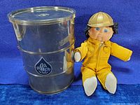 Oil Patch Kids doll in yellow coverall and hard hat seated beside a shiny metal barrel featuring the Oil Patch Kids triangular logo on a blue cloth background.
