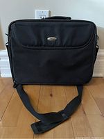 Front view of black 17-inch laptop bag with curved flap and metal logo plate, padded handle and adjustable shoulder strap on wooden floor