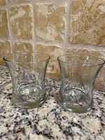 Two clear glass serving pieces with slightly flared tops photographed on granite countertop against a textured stone tile wall.