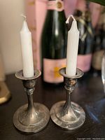 Pair of silver-toned metal candlesticks each holding a white taper candle, placed on a surface with bottles in background
