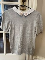 Full view of the light grey short sleeve knit shirt hanging on a black hanger, showing the white collar and overall shirt shape