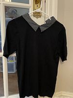 Black women's shirt hanging on white hanger showing front view with short sleeves and daisy polka dot collar in black and white pattern.