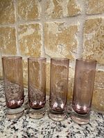 Four tall, slim vintage pink depression glass shot glasses lined up on granite countertop against textured tile wall.
